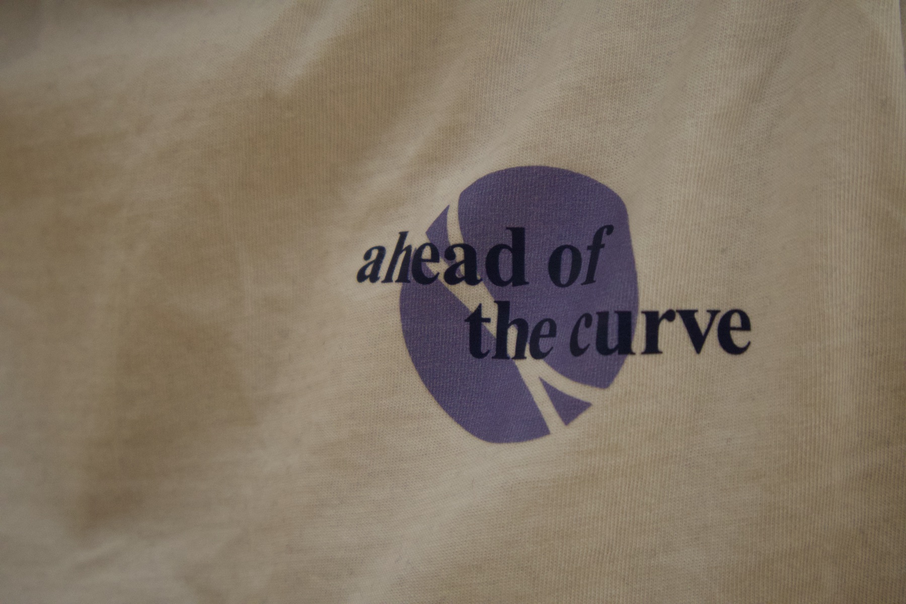 Close-up of the purple "ahead of the curve" print on a cream VSEUS tee