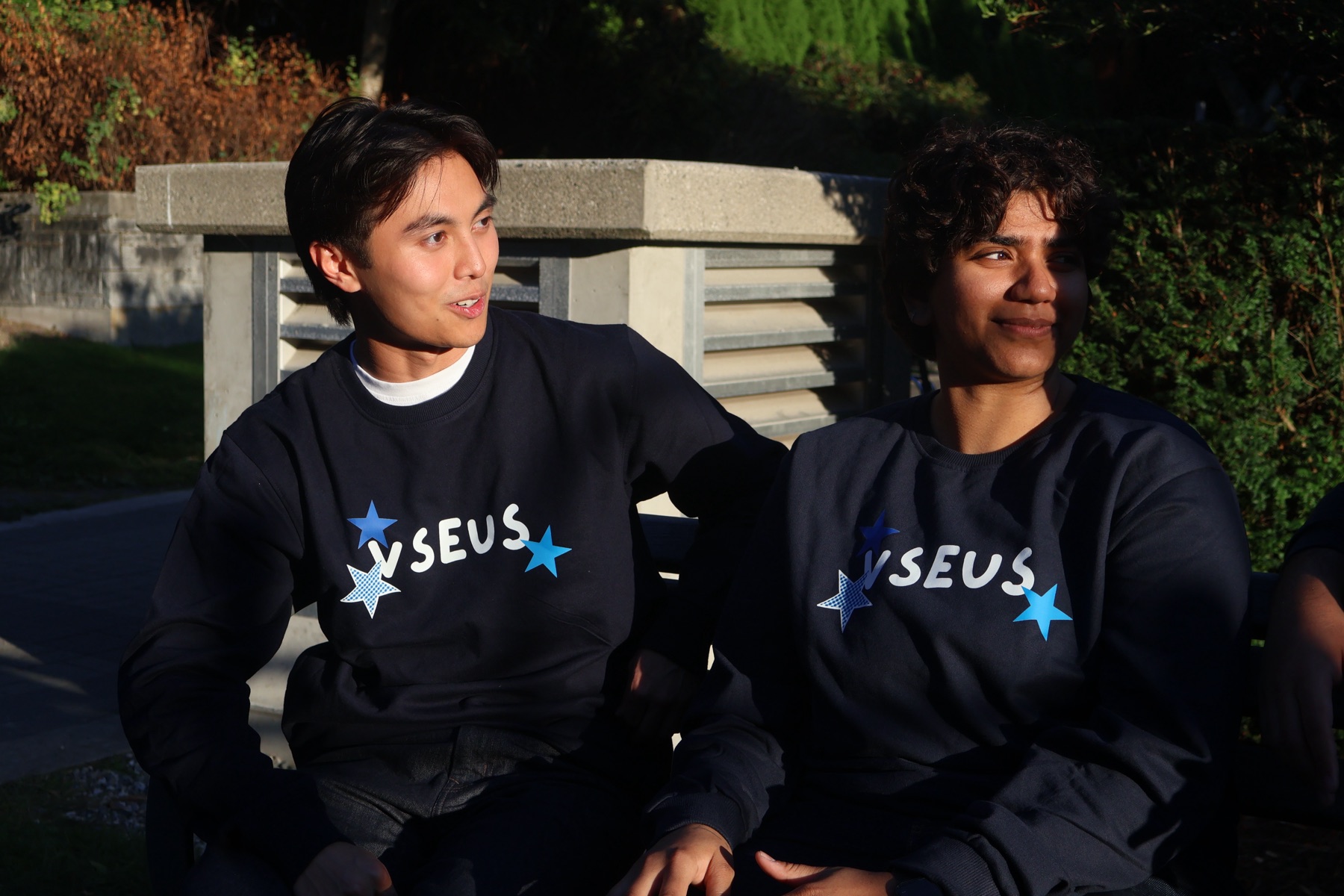 Two students on a bench in the sun wearing navy VSEUS sweaters