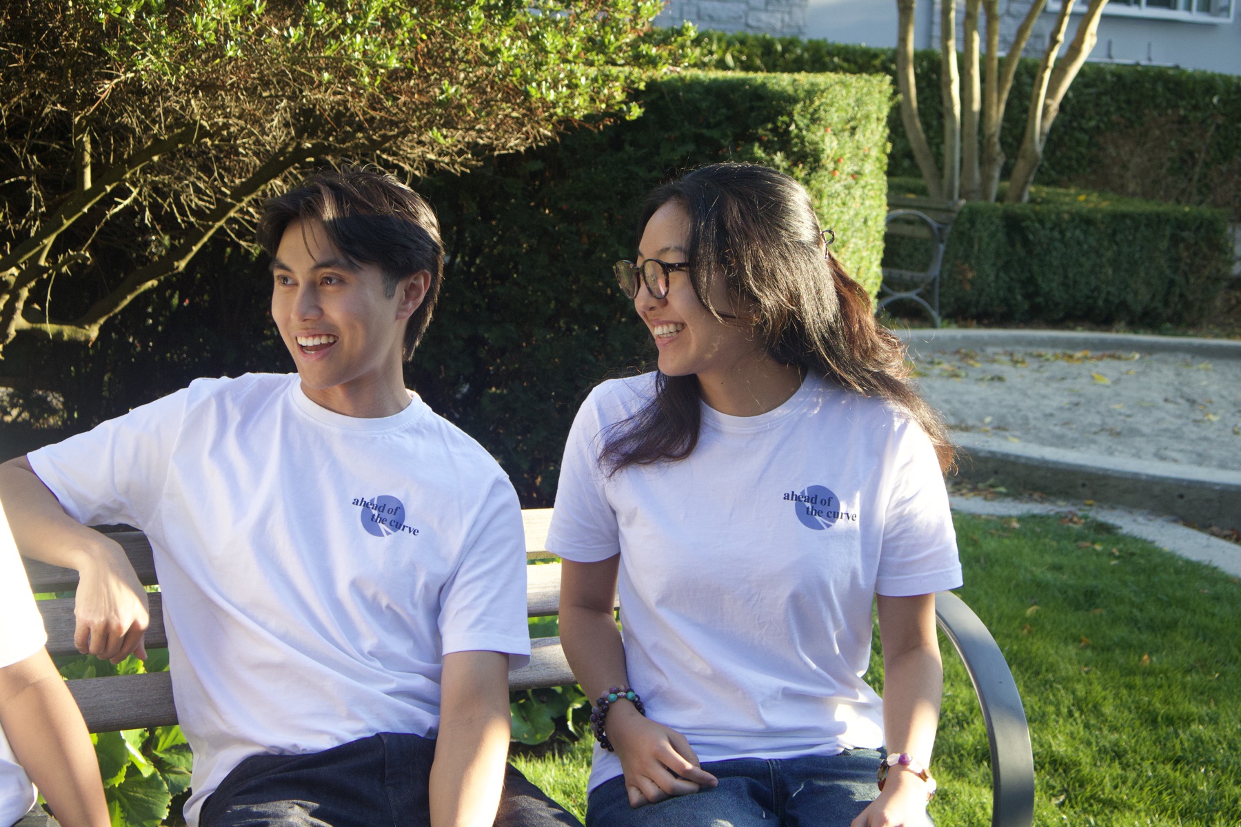 Two students on a bench outdoors in white VSEUS tees with the "ahead of the curve" print